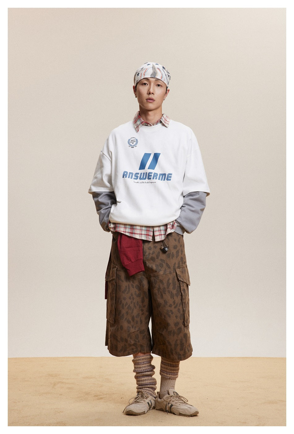 Model wearing a white graphic short-sleeve tee over gray sleeves and a plaid shirt, with patterned shorts.