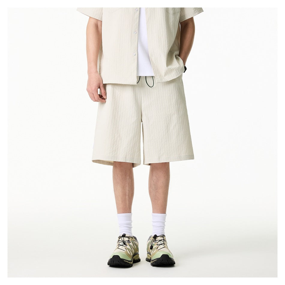 Cropped front view of a model wearing light beige knee-length shorts and a matching button-front shirt over a white tee.