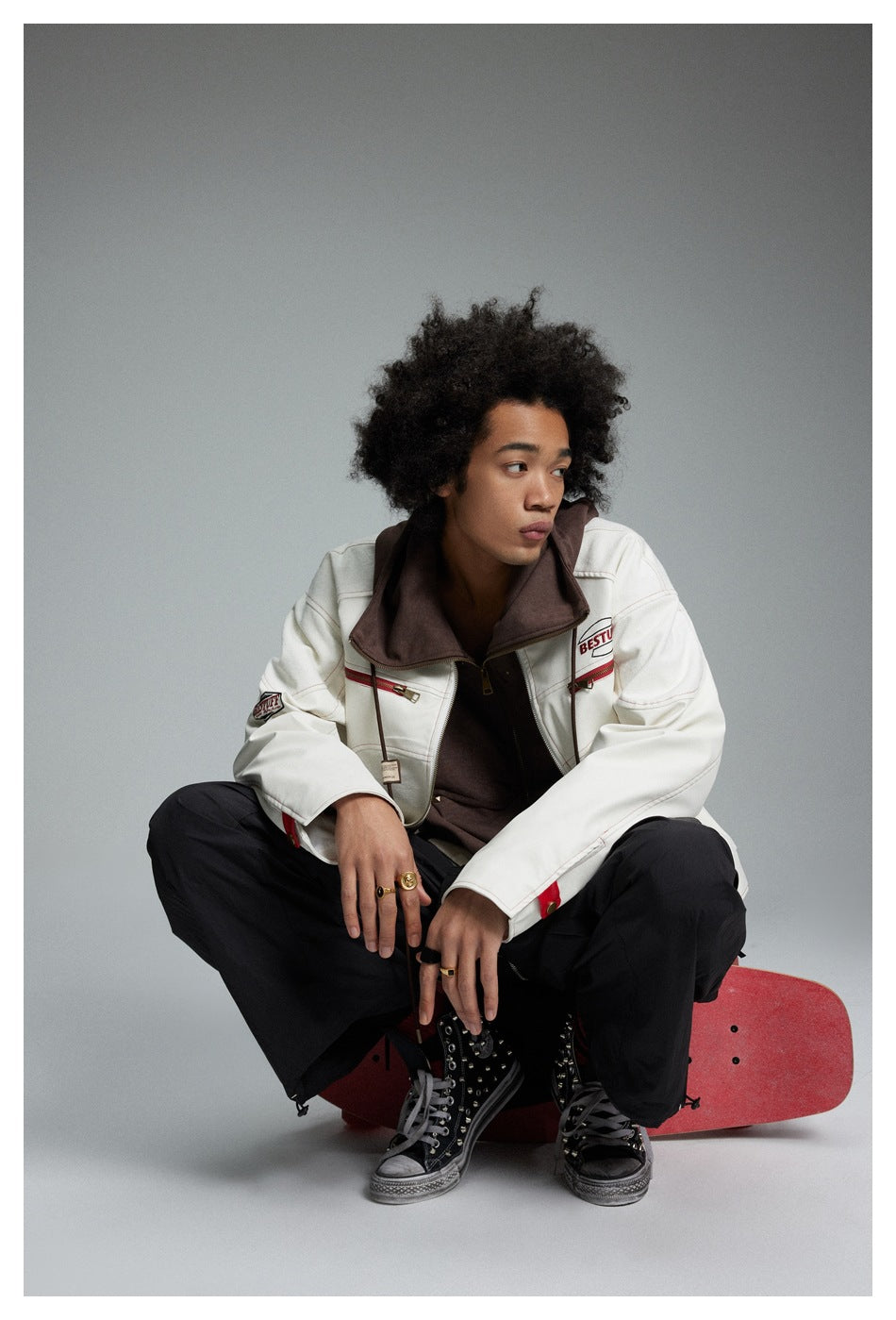 Seated model wearing a cream jacket with a brown hood and red accents, paired with loose black trousers.
