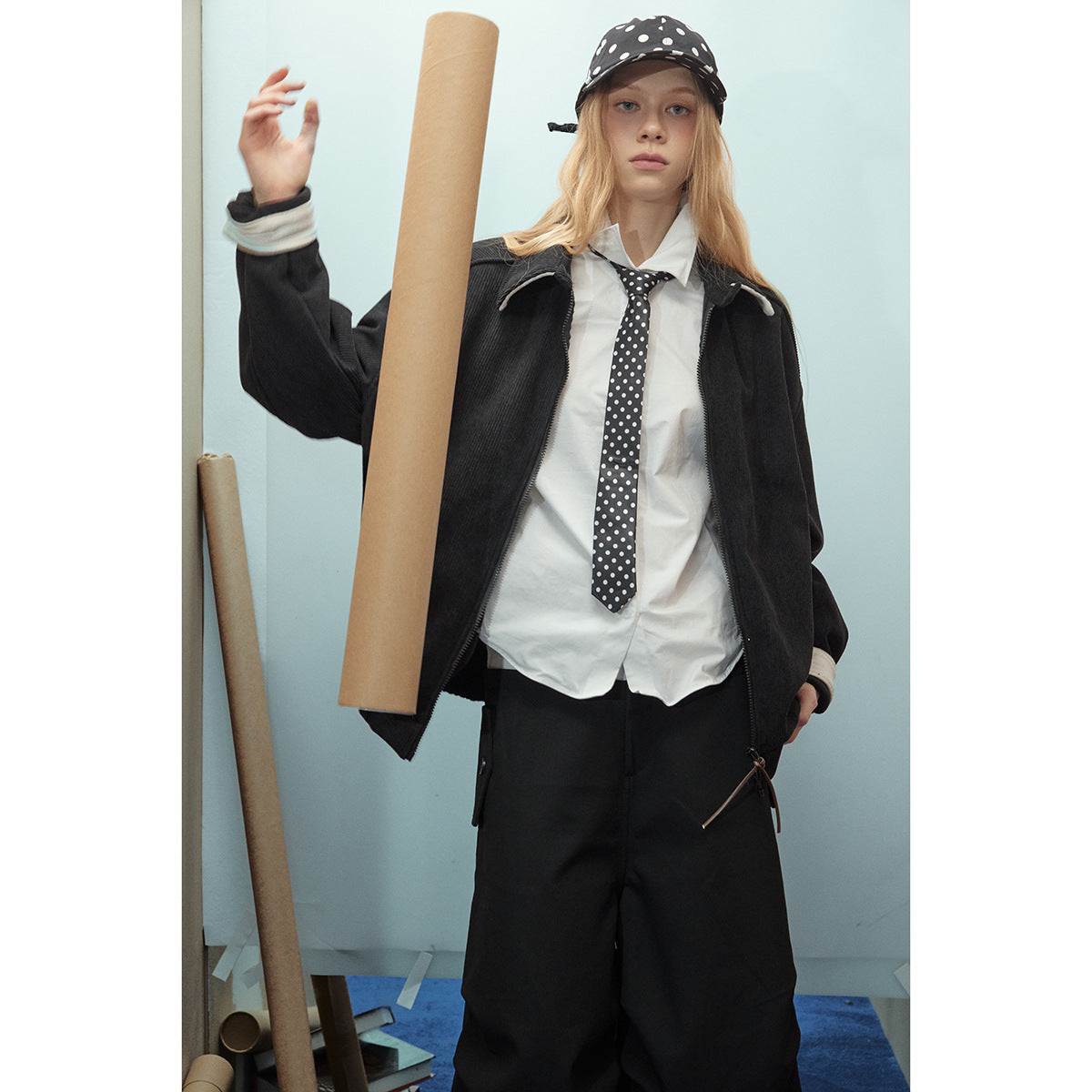 Model wearing a black zip jacket over a white shirt and polka-dot tie, with a matching cap.