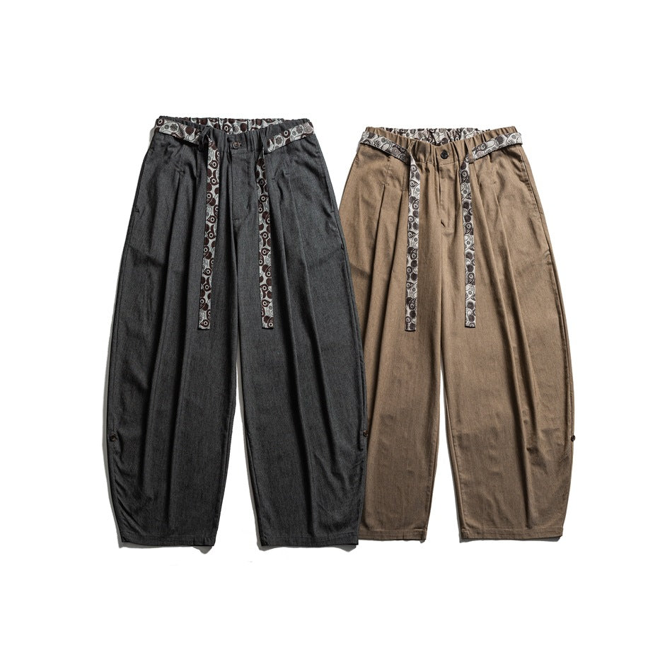 Front view of loose, tapered trousers in charcoal and tan, both with patterned waistbands and long matching straps.