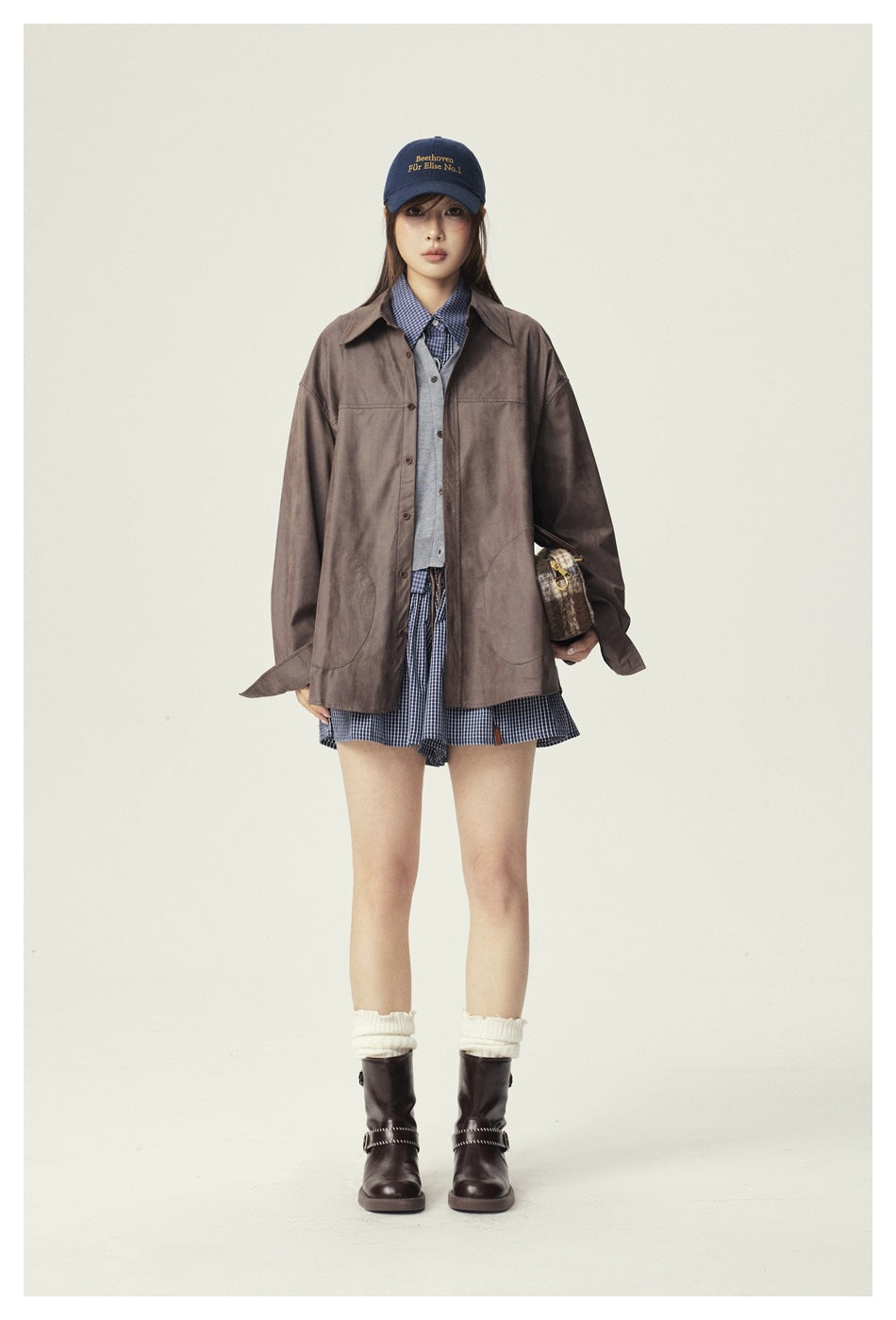 Front view of a model in an oversized brown button-up jacket over blue checked layers, with a navy cap and dark boots.