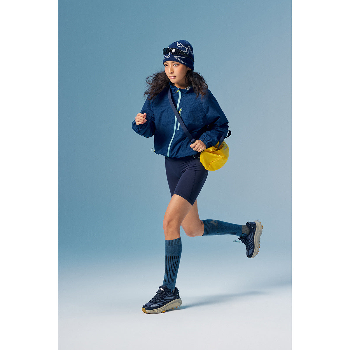 Woman running in a navy zip jacket and cycling shorts, with knee-high socks, a yellow bag, and a patterned beanie.