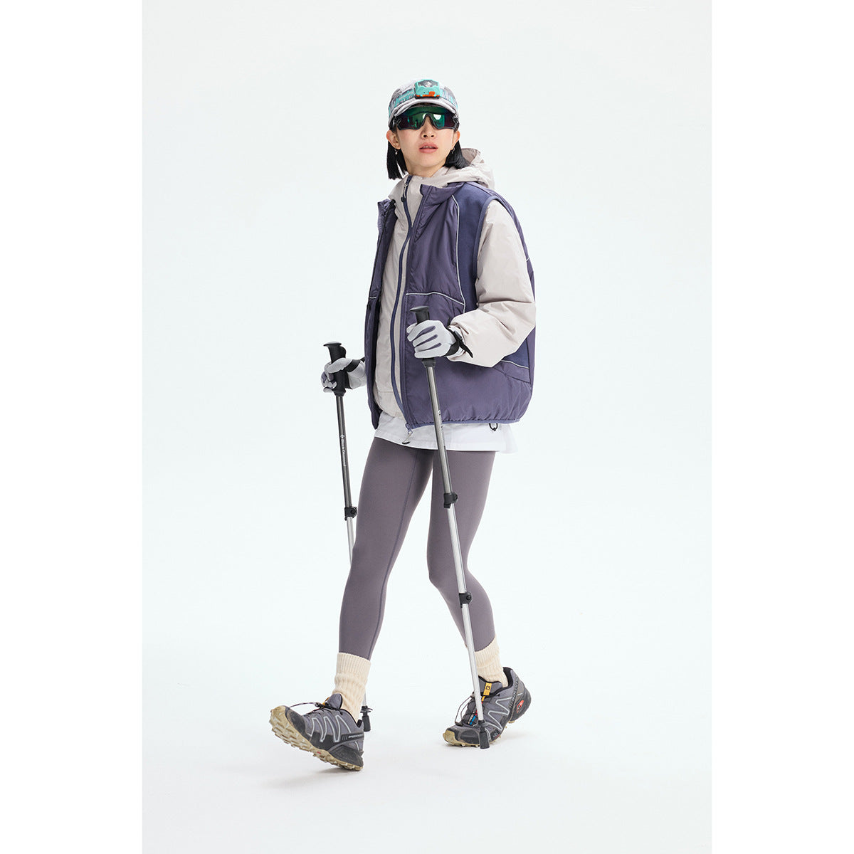Model wearing a purple sleeveless outdoor vest over a light hooded jacket, holding trekking poles.