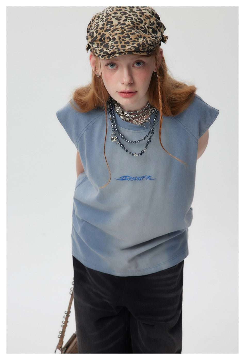Model leaning forward in a slate-blue sleeveless top with blue chest embroidery, black trousers and a leopard-print cap.
