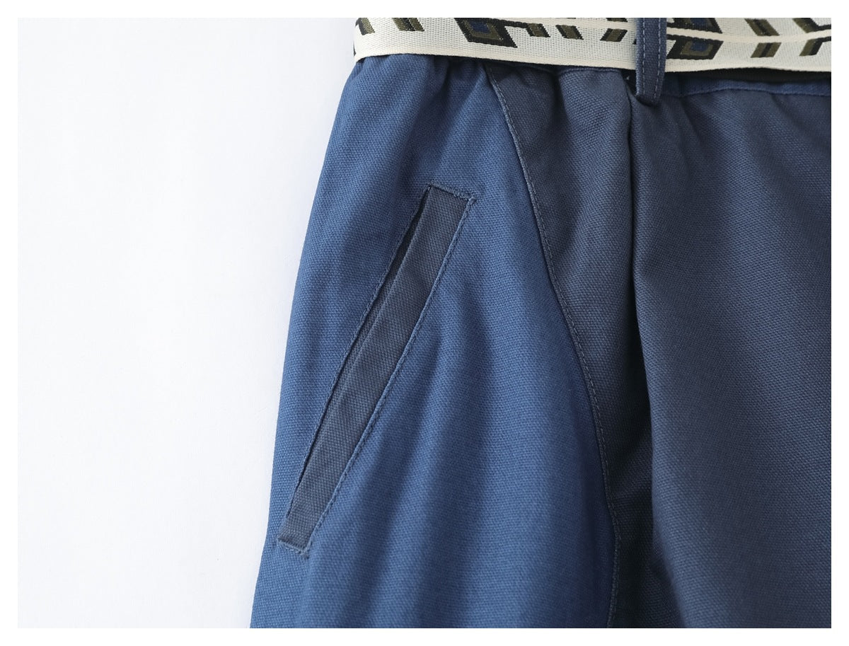 Close-up of blue trousers showing an angled front pocket and the patterned belt at the waistband.
