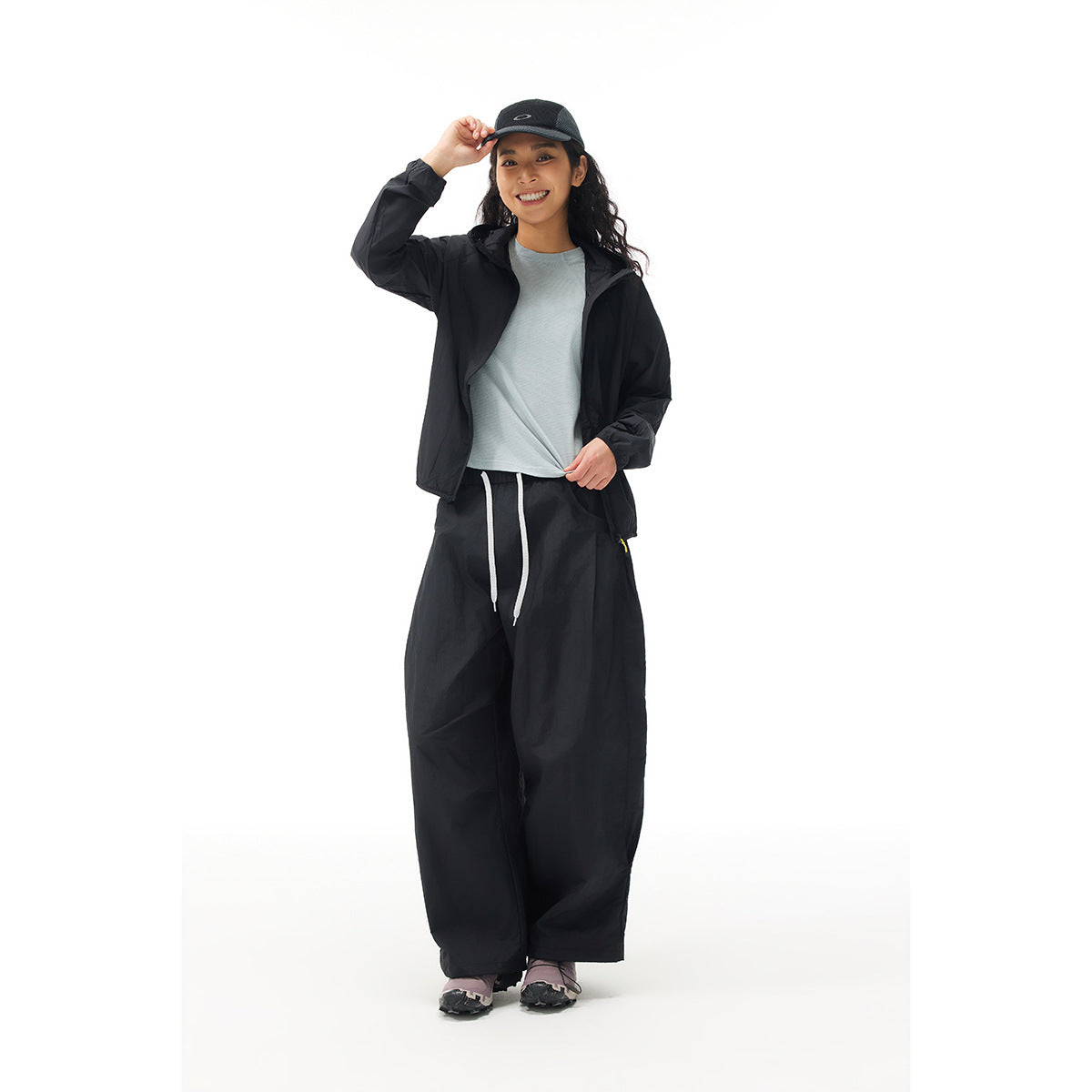 Woman wearing a black hooded windbreaker and loose black drawstring pants, with a gray shirt and cap.