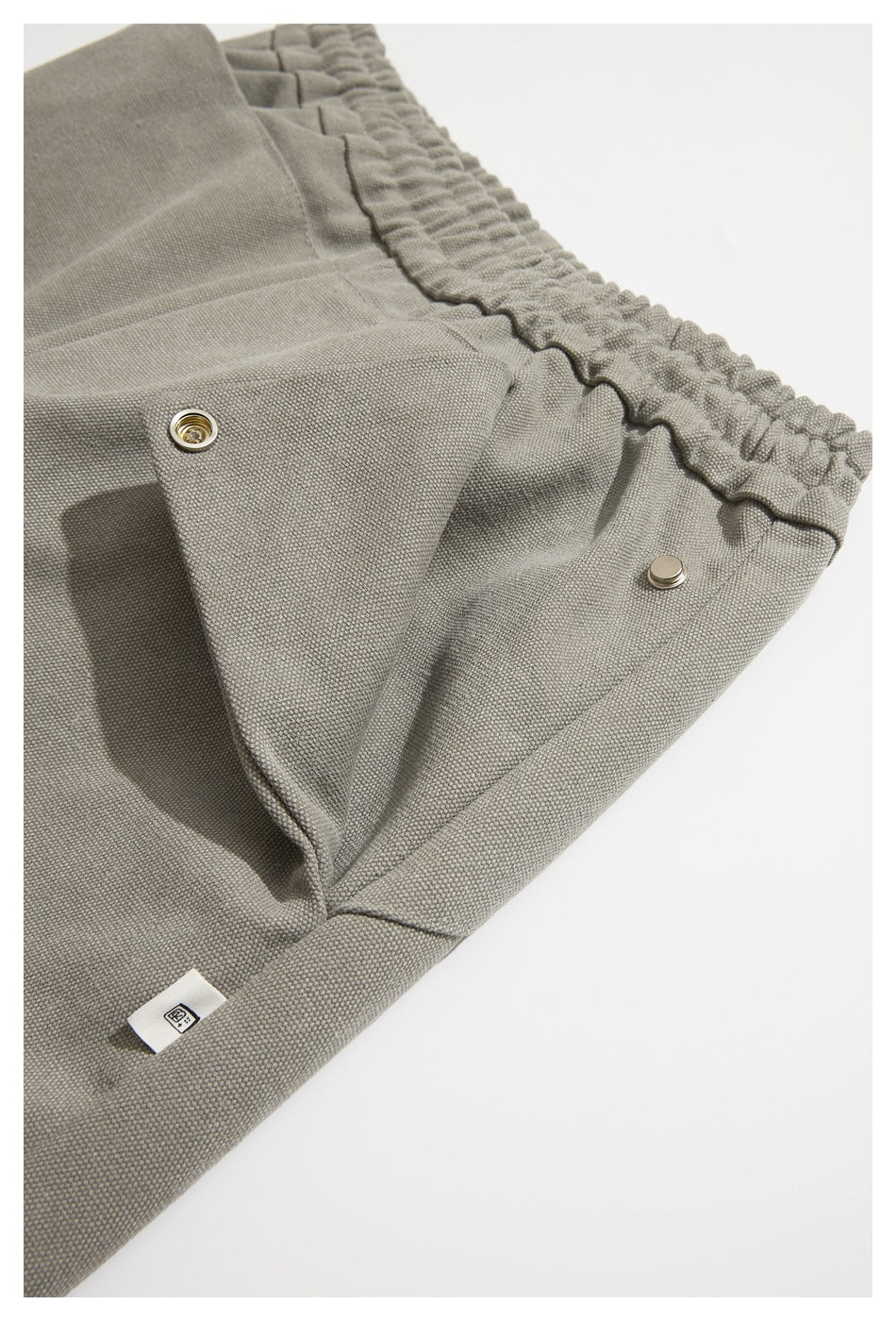 Close-up of taupe-gray trousers showing a gathered elastic waistband, snap fastening and a small fabric tag.