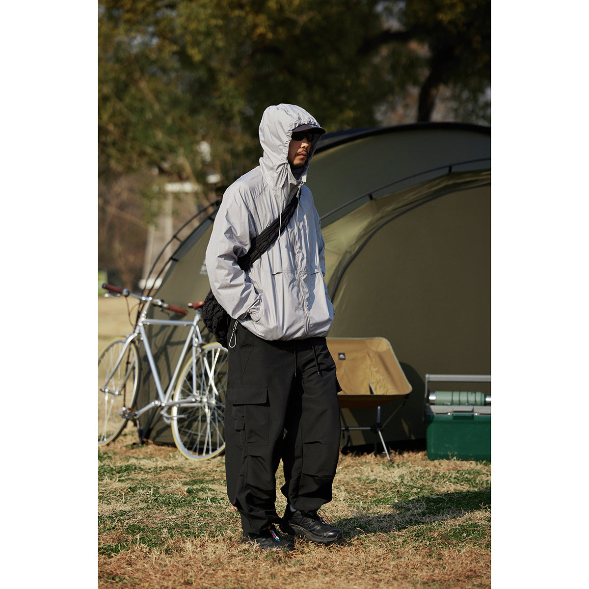 Model wearing a light gray hooded zip jacket and black cargo pants at a campsite.