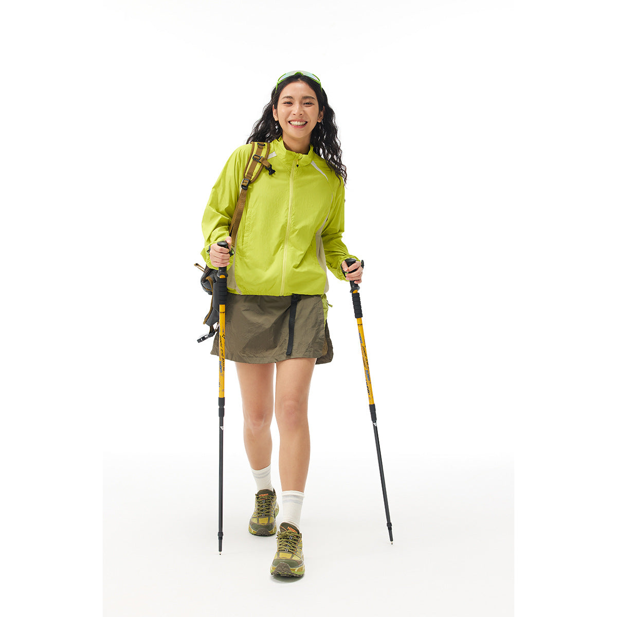 Woman wearing a lime-green zip jacket and olive hiking skirt, standing with trekking poles against a white background.