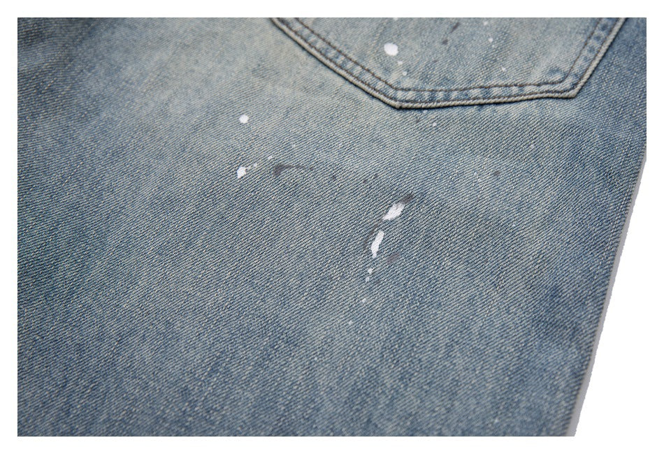 Close-up of light blue denim with scattered white paint-like marks below a pocket.