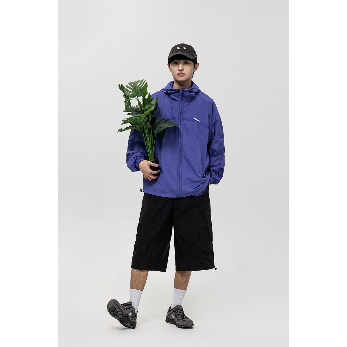 Front view of a model wearing a blue-violet hooded zip jacket, holding a leafy plant.
