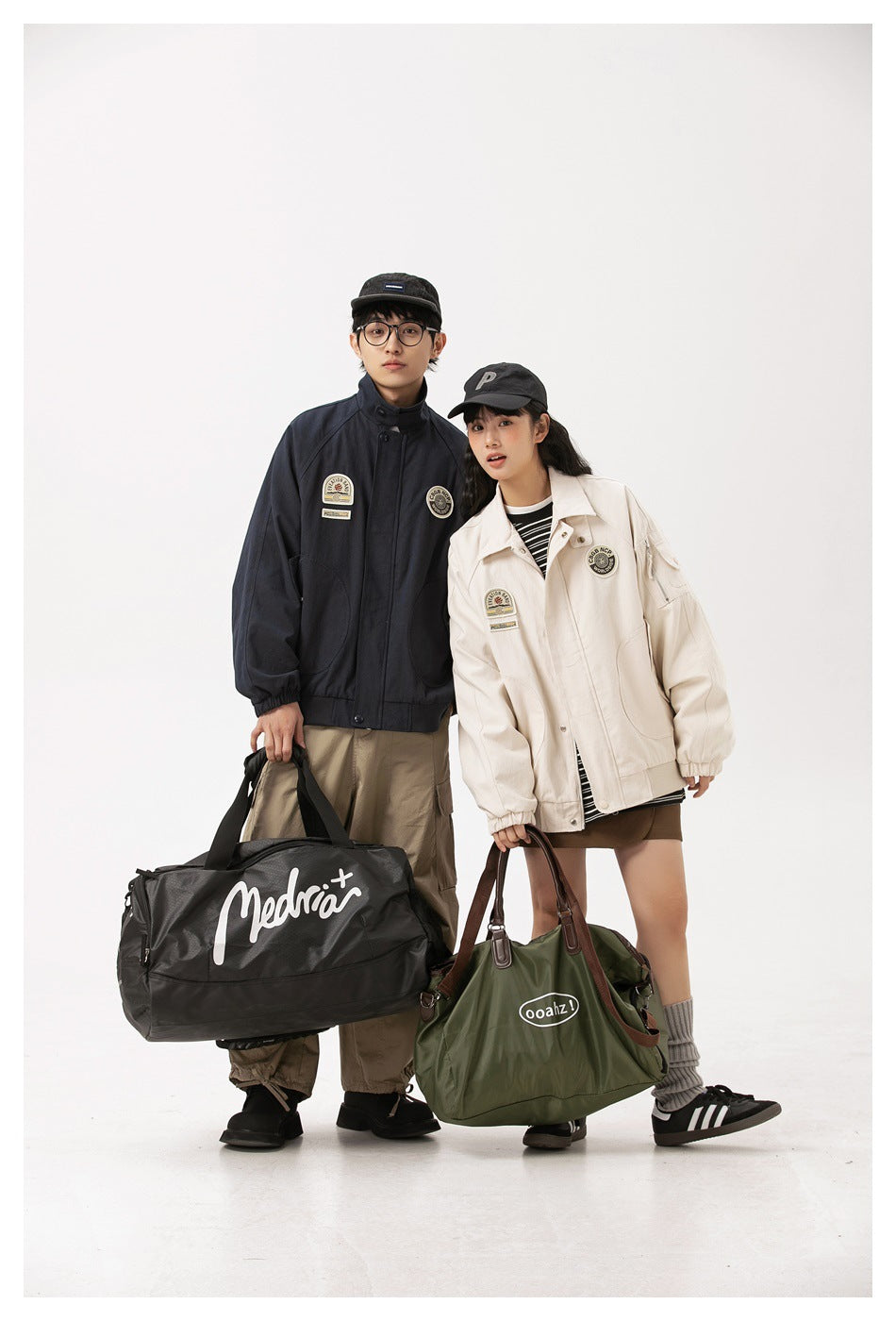 Two models wear dark navy and cream patch jackets, carrying large duffel bags against a plain studio backdrop.