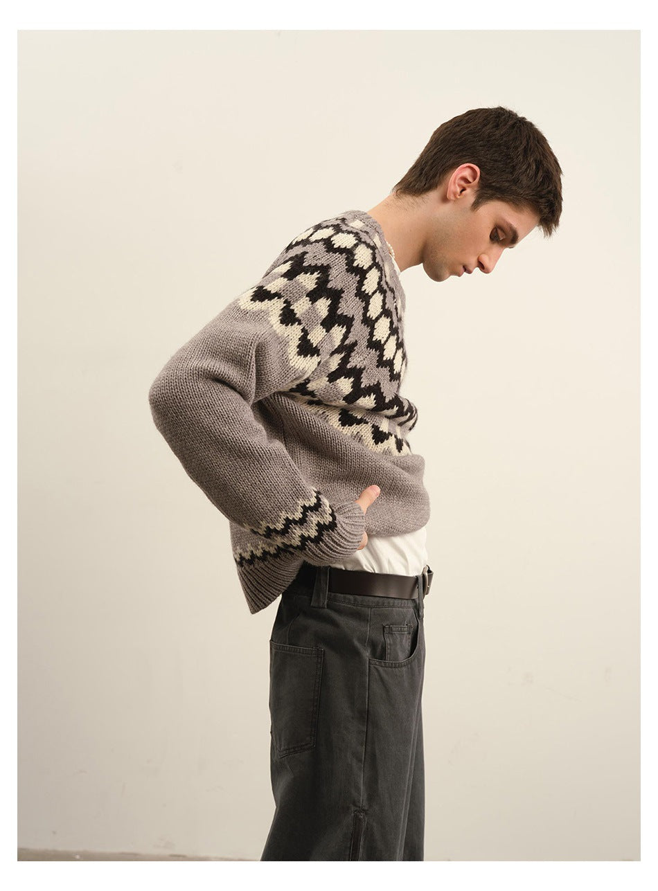 Side view of model in gray patterned knit sweater showing sleeve, ribbed cuff and zigzag pattern, with dark pants and belt