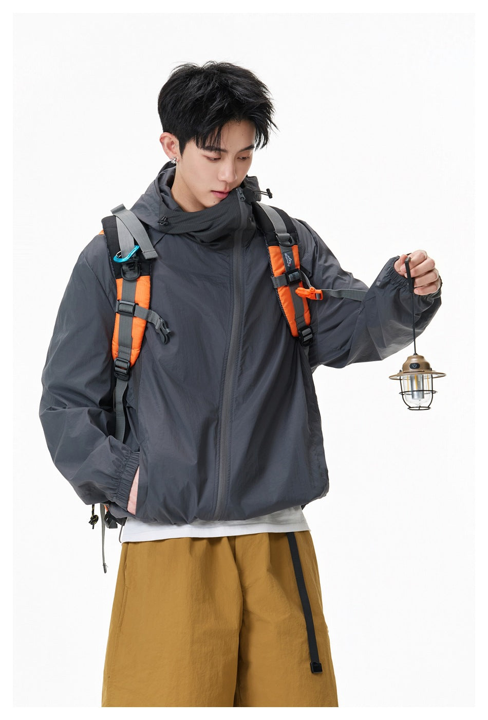 Model wearing a charcoal hooded jacket with orange backpack straps, holding a small lantern.