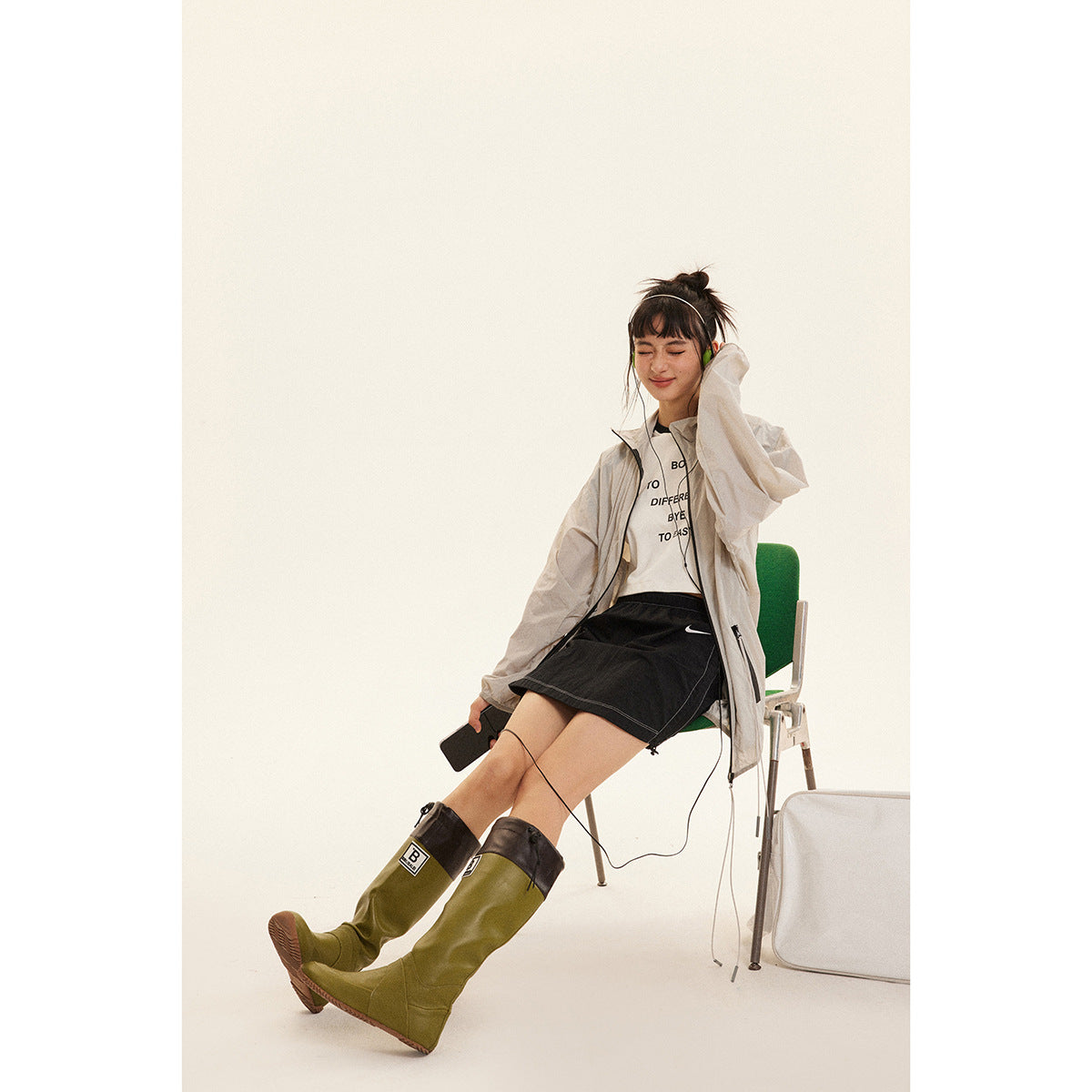 Seated model wearing a light gray zip jacket, white graphic tee, black skirt and olive-green knee-high boots.