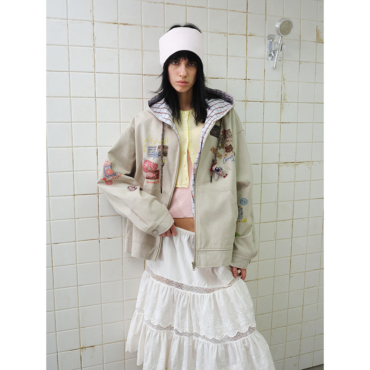 Model wearing a taupe zip hoodie with colorful graphic patches and a plaid-lined hood over a white tiered skirt.