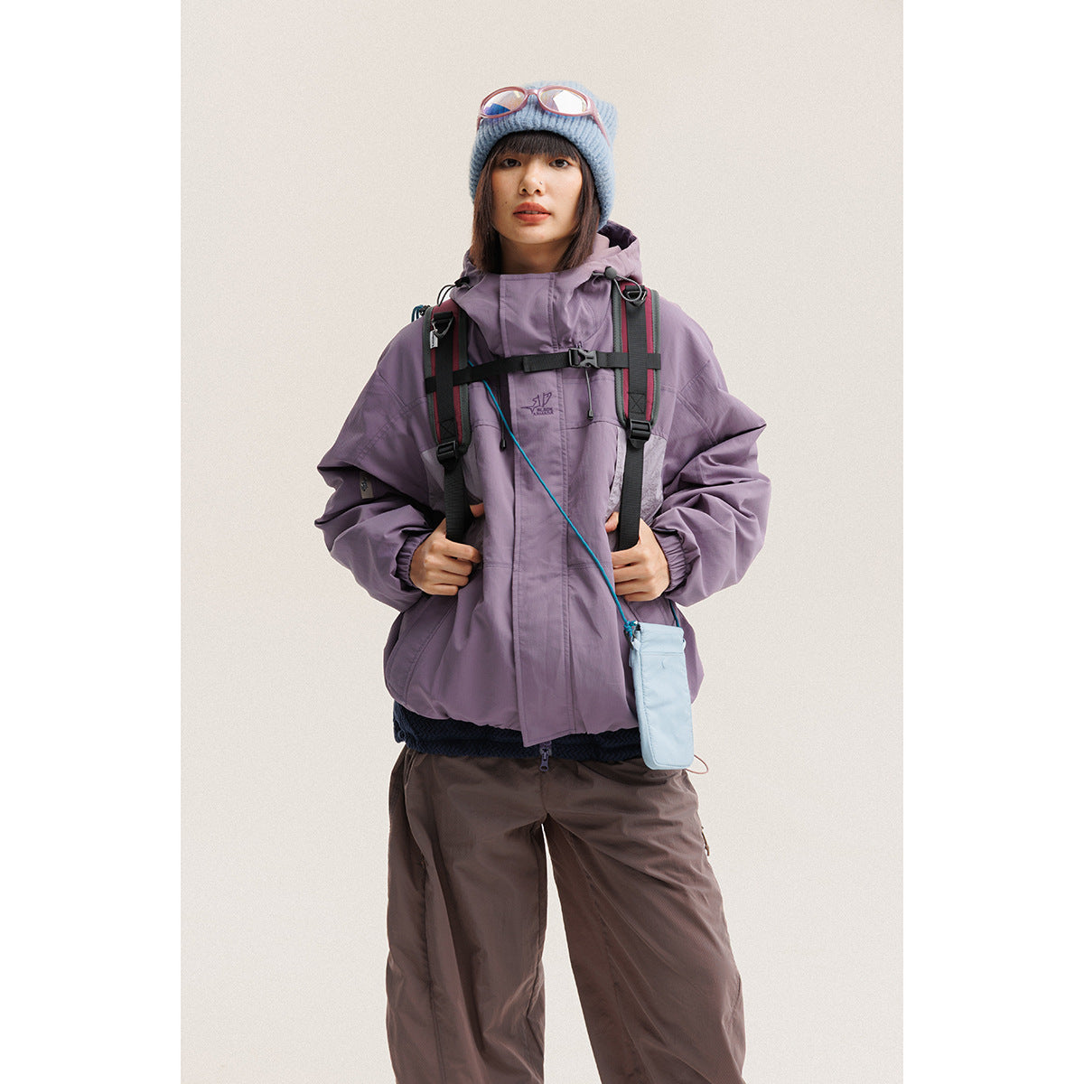 Model wearing a lavender hooded jacket with a black backpack, blue beanie and brown pants, facing forward.