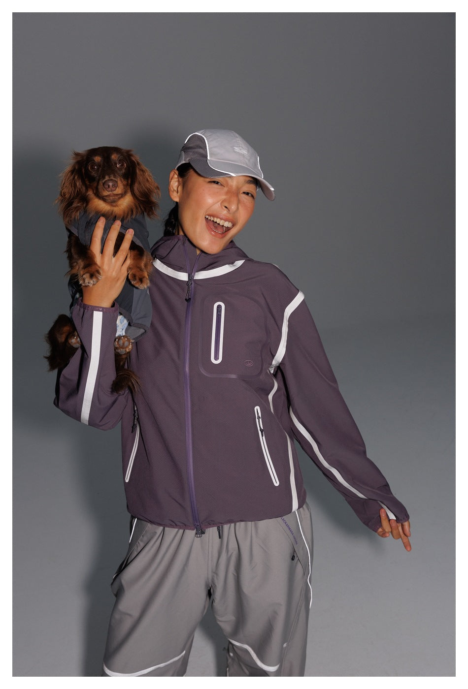 Model holding a small dog and wearing a dark purple jacket with white trim and gray pants.