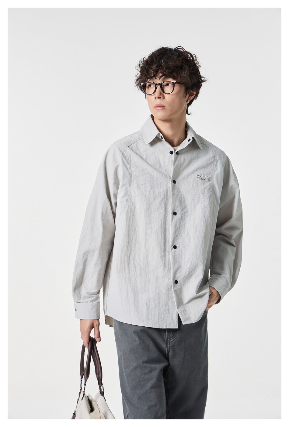 Man in glasses wearing a light gray button-front shirt and gray pants while holding a tote bag
