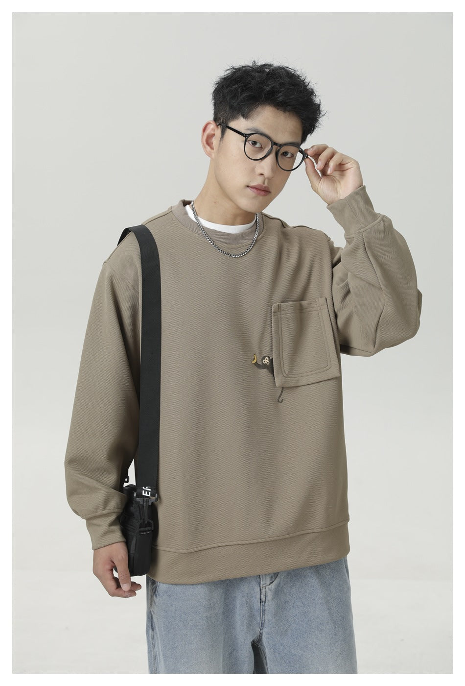 Model wearing a taupe crewneck sweatshirt with a chest pocket and small embroidered motif, paired with light jeans.