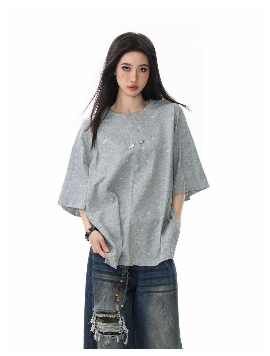 Front view of a model in a loose light-gray T-shirt with scattered pale marks and a small chest emblem.