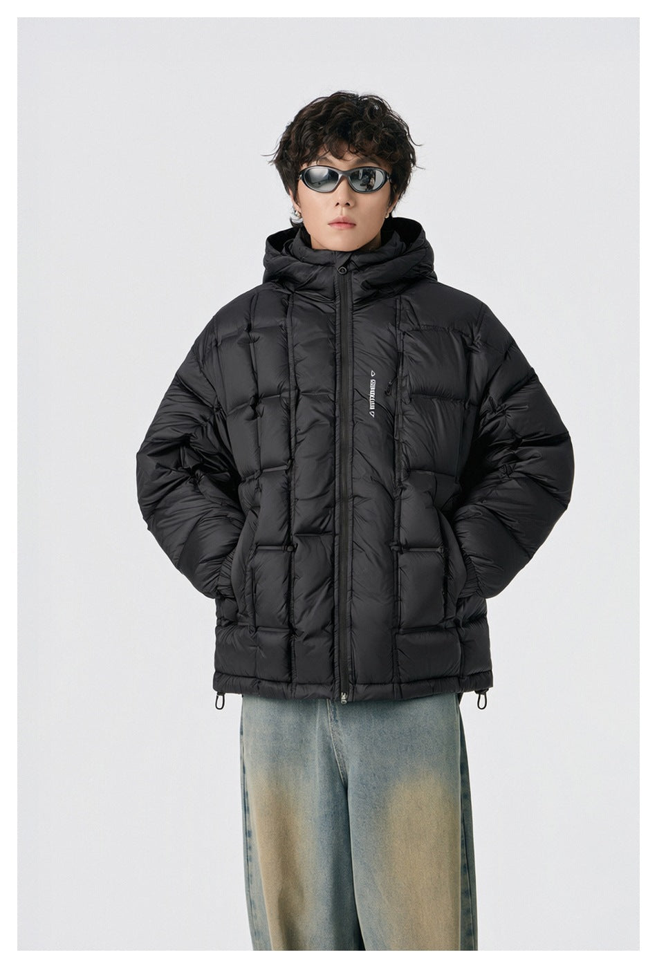 Front view of a model wearing a black hooded puffer jacket with a front zipper, quilted panels, and jeans.