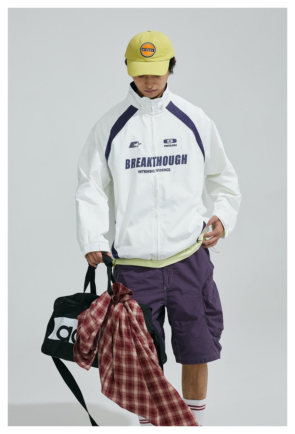 Model wearing a white zip-up jacket with navy panels and English lettering, paired with purple shorts and a yellow cap.