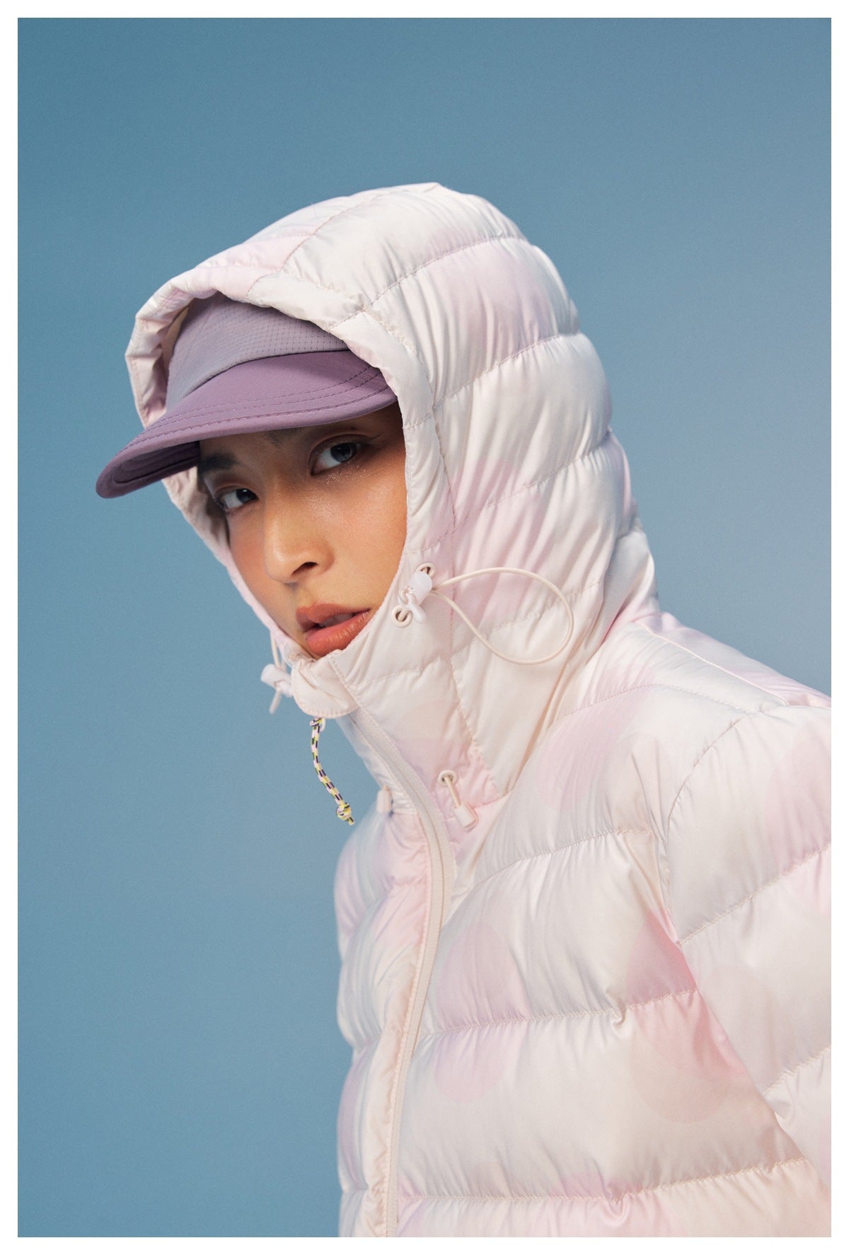 Close-up of a model wearing a pale pink quilted hooded jacket and mauve cap, showing the hood and drawcord details.