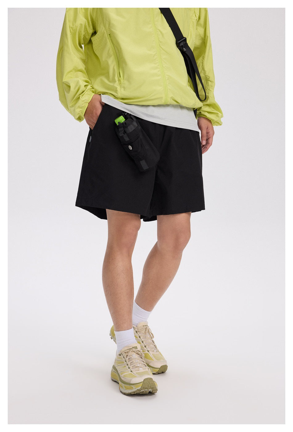 Angled view of a model wearing black shorts with an attached utility pouch, a lime jacket, and beige sneakers.