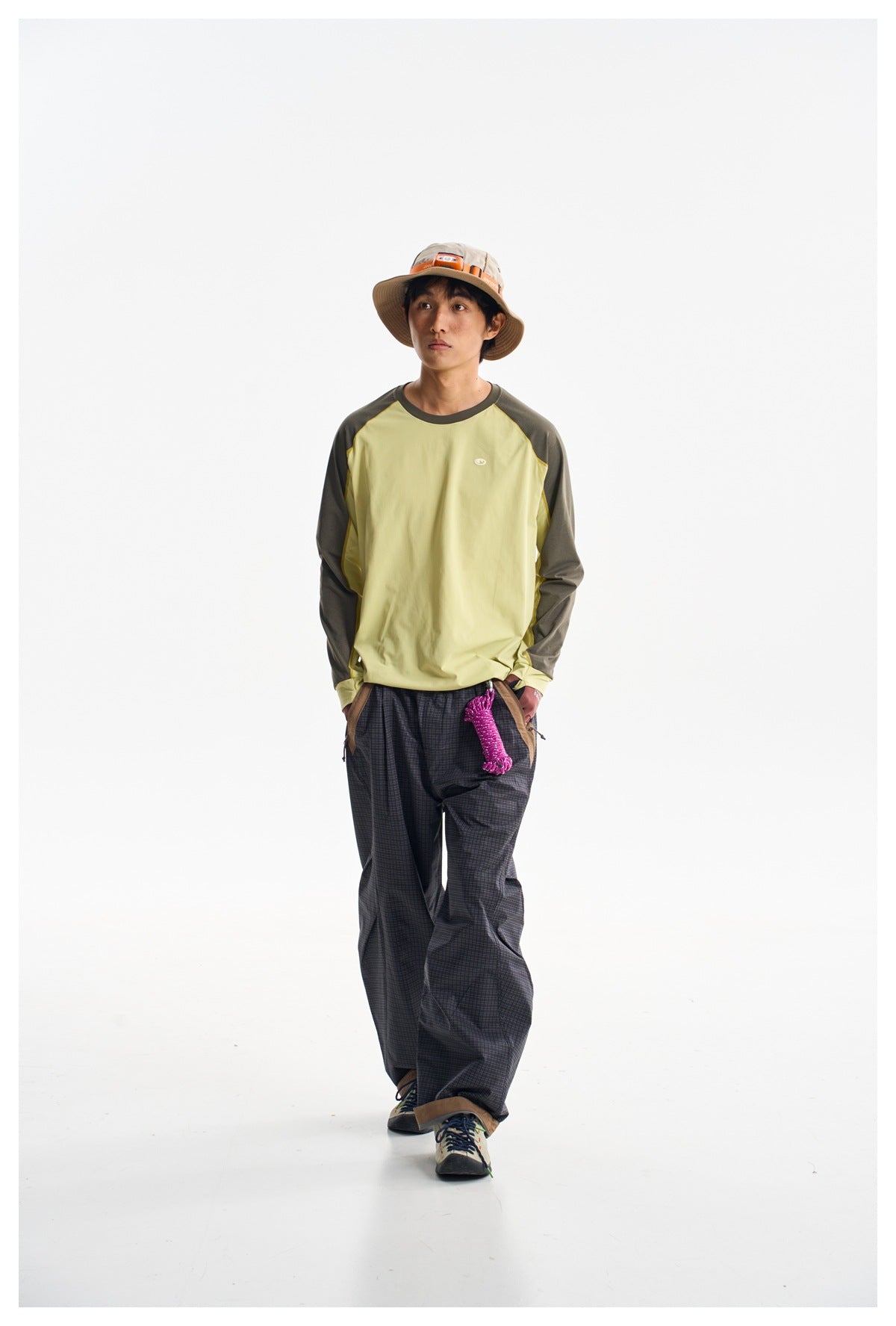 Model walking in a pale yellow and dark olive raglan shirt with loose charcoal plaid pants.