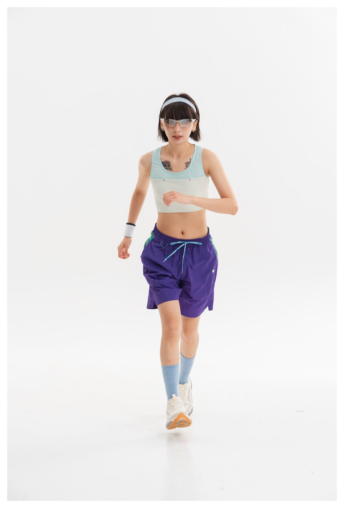 Model walking in a mint and ivory cropped tank, purple shorts, blue socks, and white sneakers.