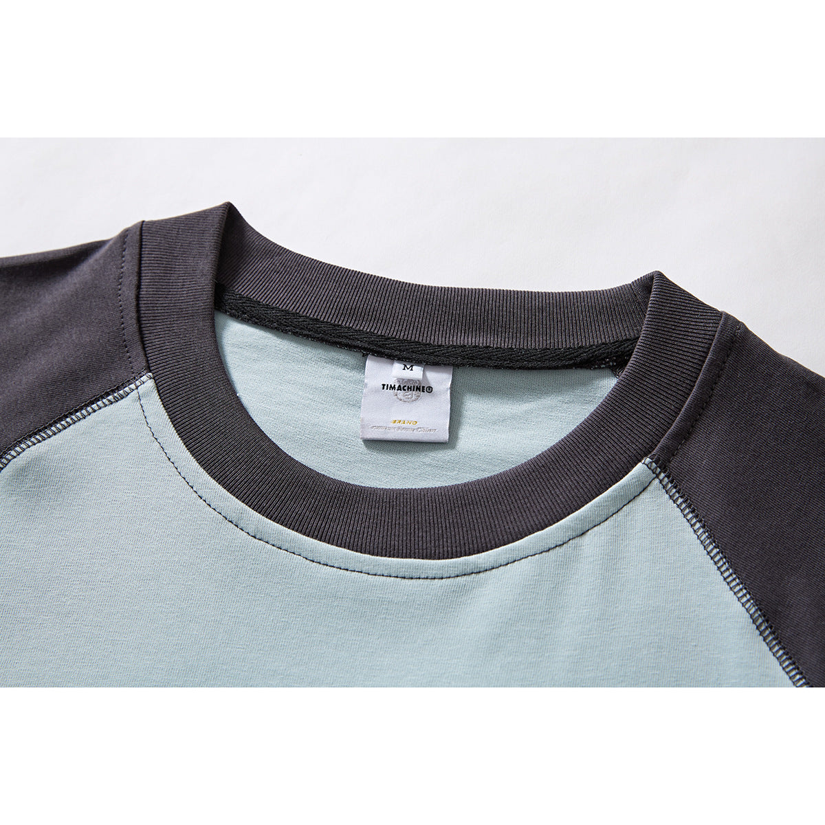 Close-up of a pale blue and charcoal raglan T-shirt neckline, ribbed collar, seams, and sewn neck label.