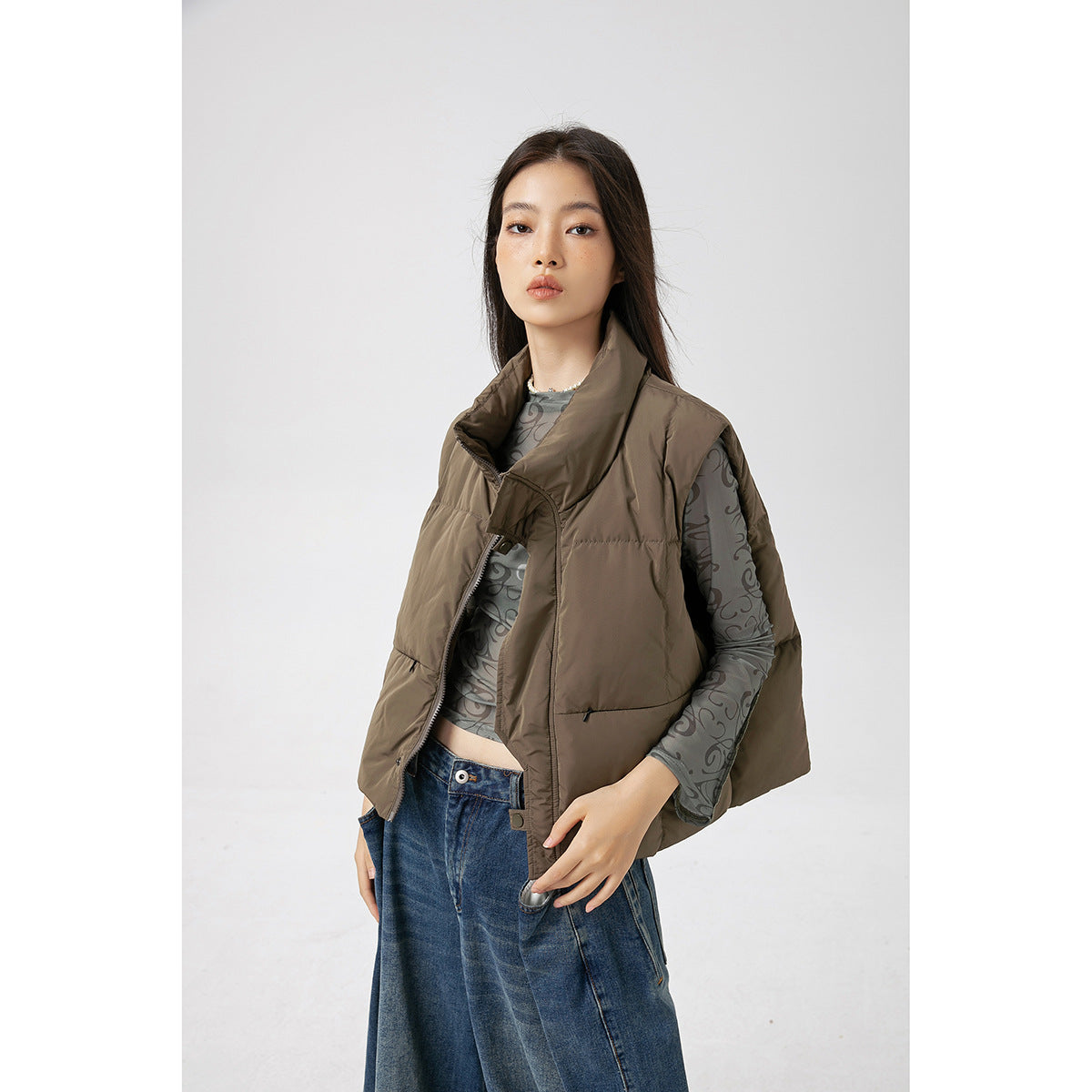 Model wearing a taupe padded vest with a high collar over a gray patterned sheer top and wide-leg blue jeans.