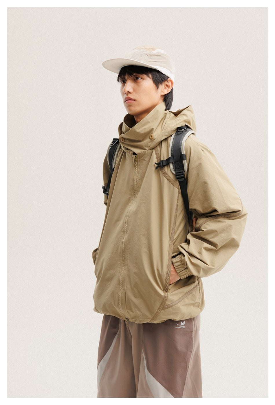 Closer view of a model in a tan hooded zip jacket with a high buttoned collar and taupe color-block pants.
