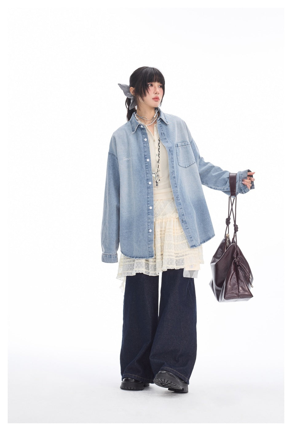 Full-length model in a light blue oversized denim shirt over cream lace layers and dark wide-leg jeans, carrying a burgundy