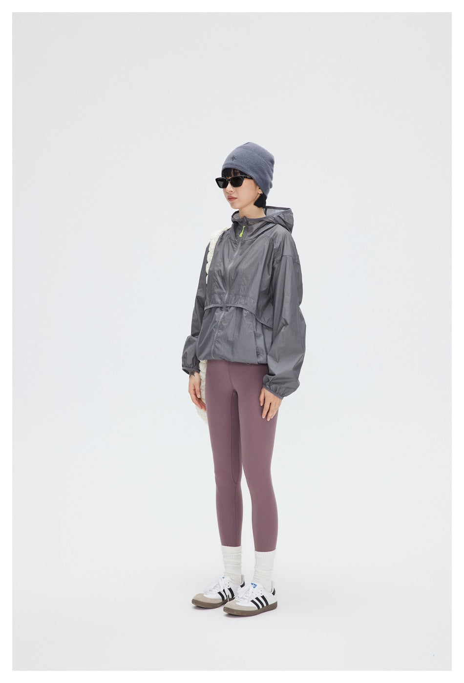 Model wearing a gray hooded windbreaker with mauve leggings, a blue-gray beanie and sunglasses, standing in a studio.