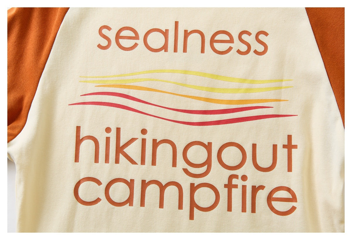 Close-up of a cream shirt with rust-orange sleeves and rust lettering reading “sealness hikingout campfire,” above colored