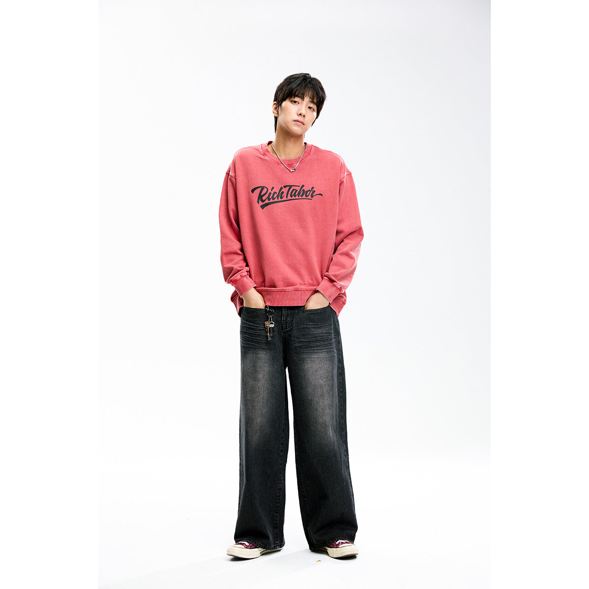 Model wearing a coral-pink crewneck sweatshirt with dark script print and loose black jeans.
