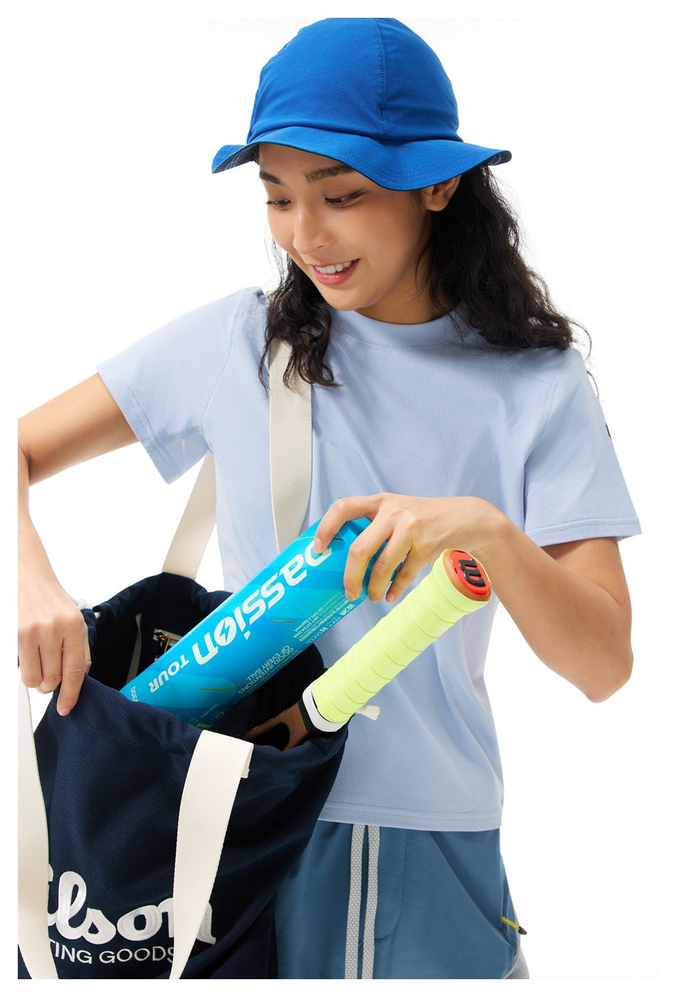 Woman in a light blue T-shirt and blue bucket hat placing a ball can and racket into a tote bag