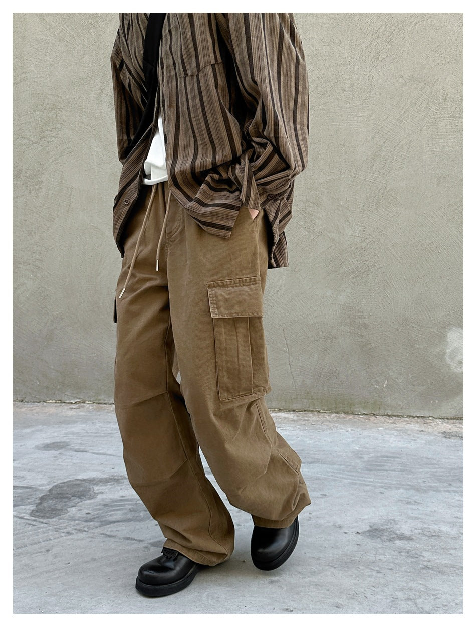 Angled view of loose brown cargo pants with a drawstring waist and a large thigh pocket, worn with a striped shirt.