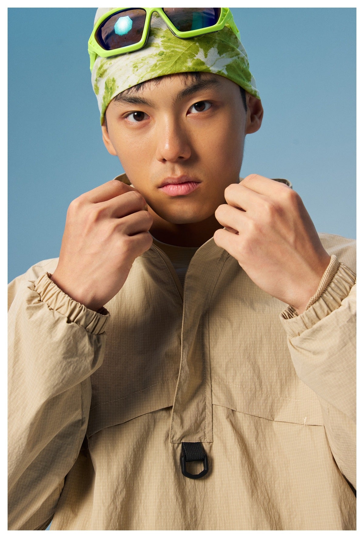 Close-up of a model adjusting the collar of a beige pullover jacket, showing its front flap and black loop.
