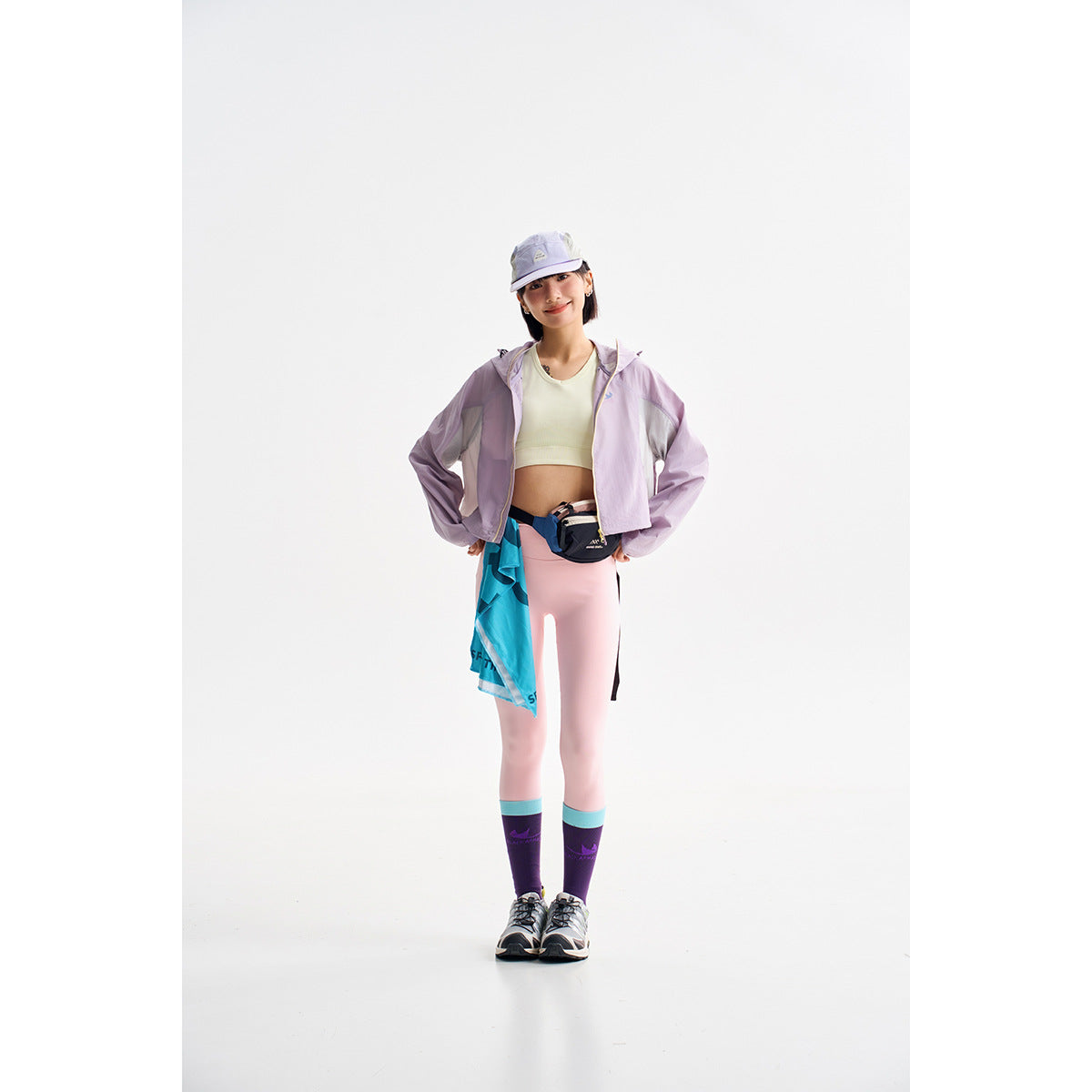 Model wearing a cropped lavender color-block hooded jacket with pink leggings, a white top, cap and running accessories.