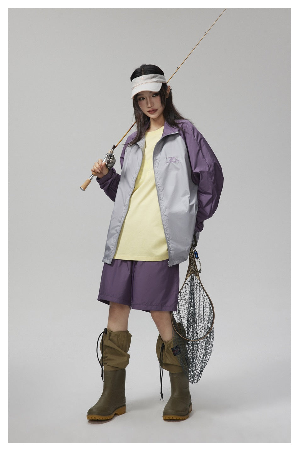 Model wearing purple shorts with a gray and purple jacket, yellow tee, visor, and rubber boots, holding a fishing rod