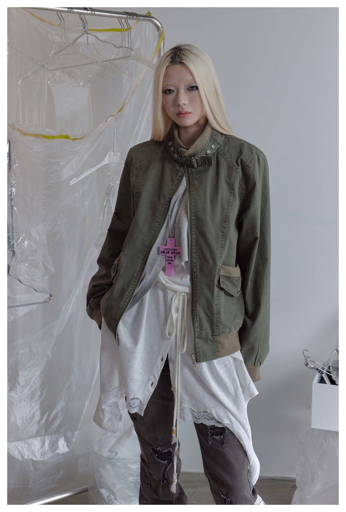 Model wearing an olive zip-front jacket with a high buckled collar and flap pockets over a white layered outfit.