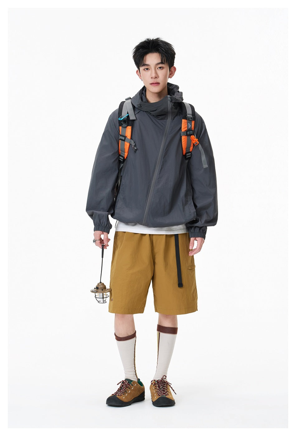 Full-length model wearing a charcoal hooded jacket with orange backpack straps and mustard shorts.