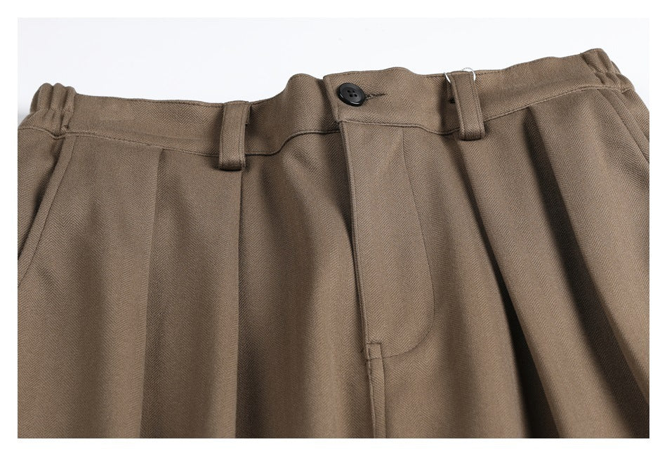 Close-up of taupe-brown trousers showing the waistband, belt loops, button, pleats, and fabric texture.