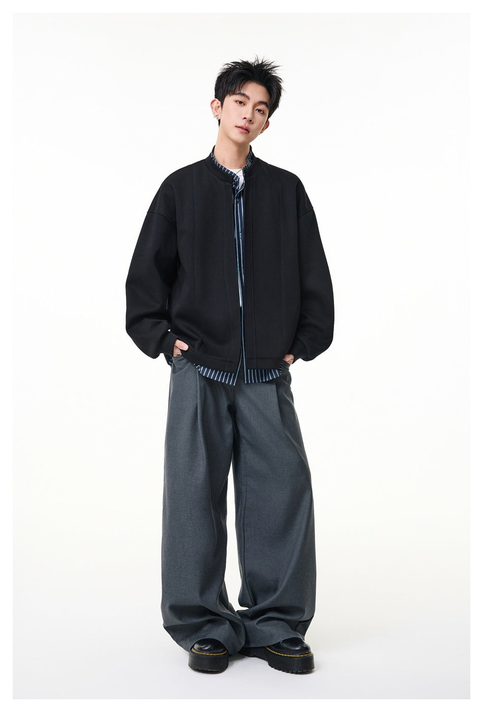 Full-length model wearing an oversized black stand-collar jacket over a blue striped shirt and wide gray trousers.