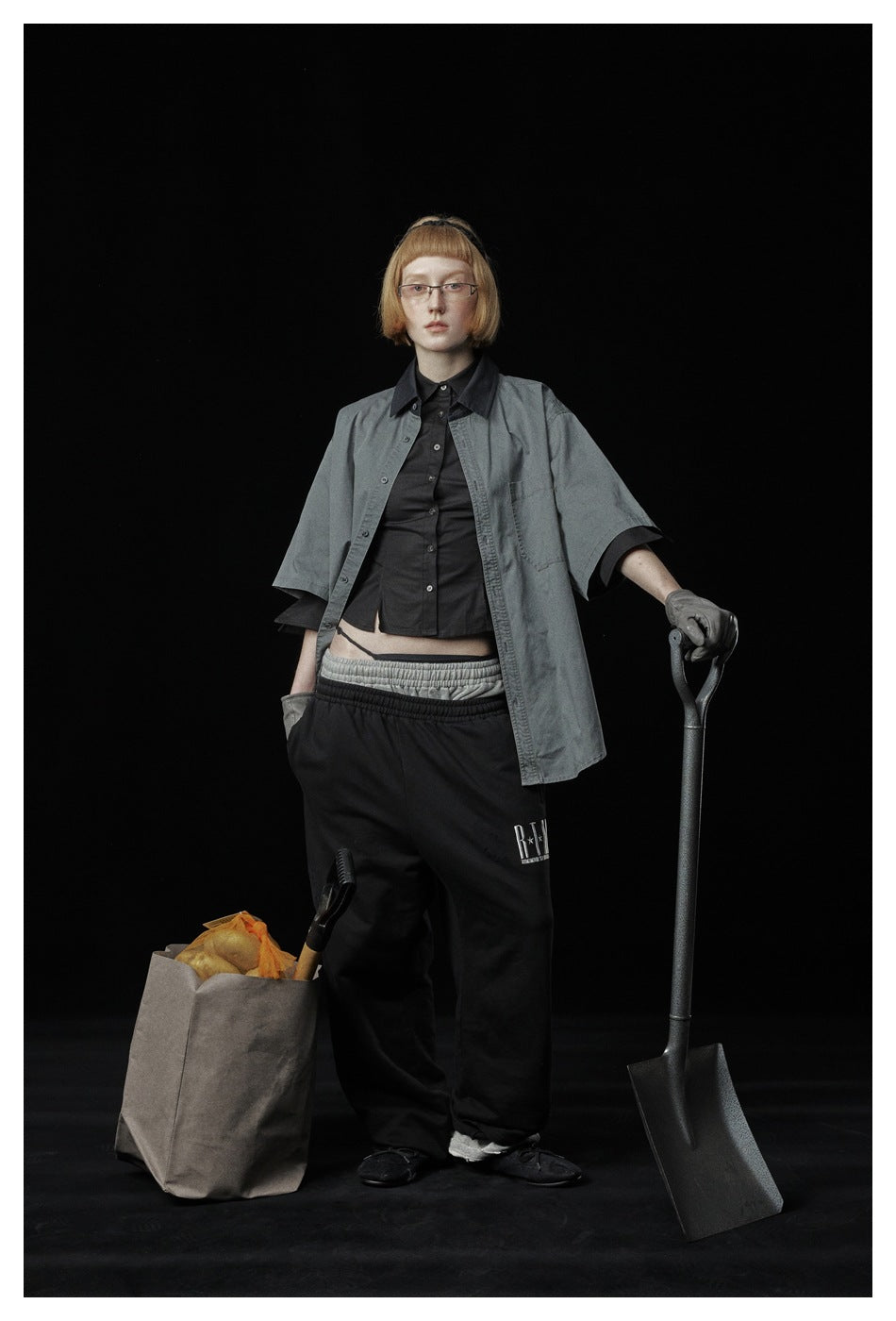Person wearing the gray short-sleeve shirt open over a black shirt and black sweatpants, holding a shovel