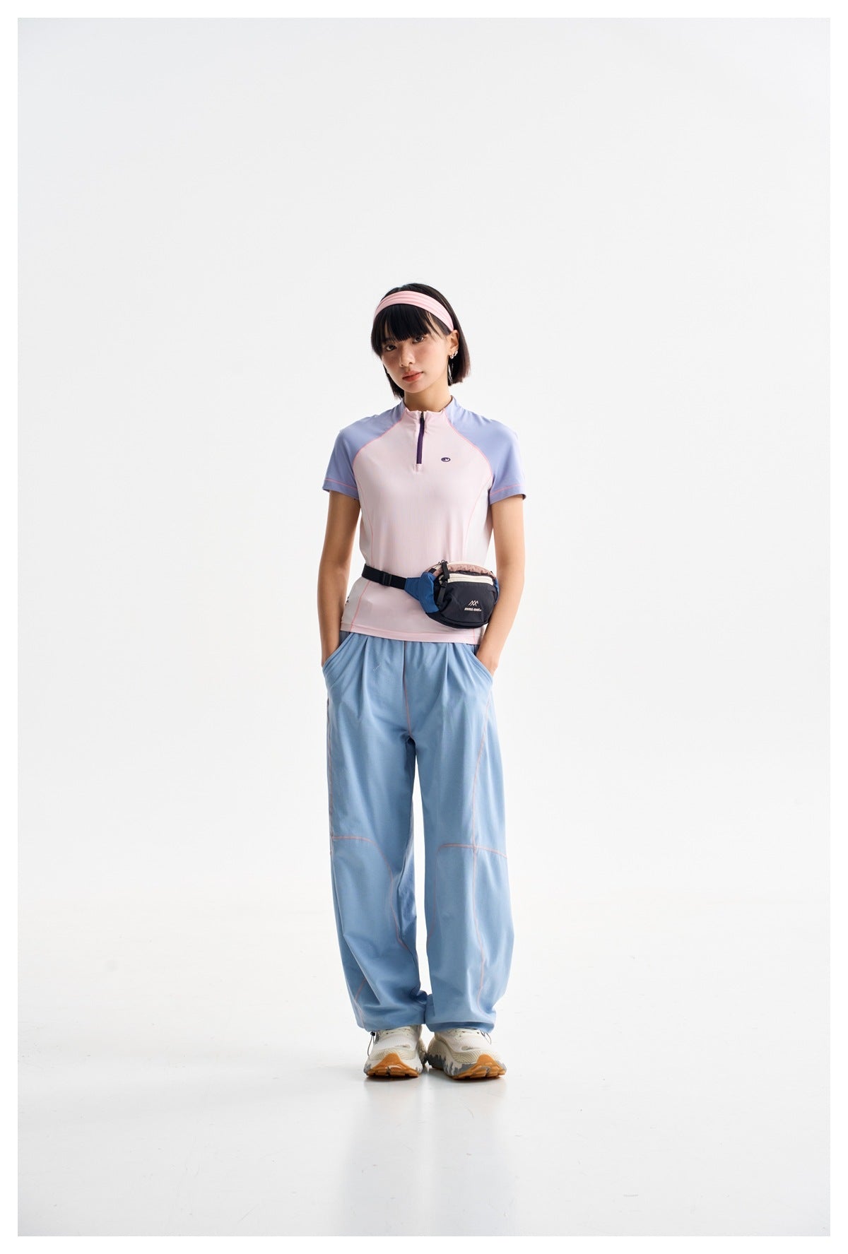 Model wearing a pale pink and blue zip-neck top with loose blue trousers and a waist pack.